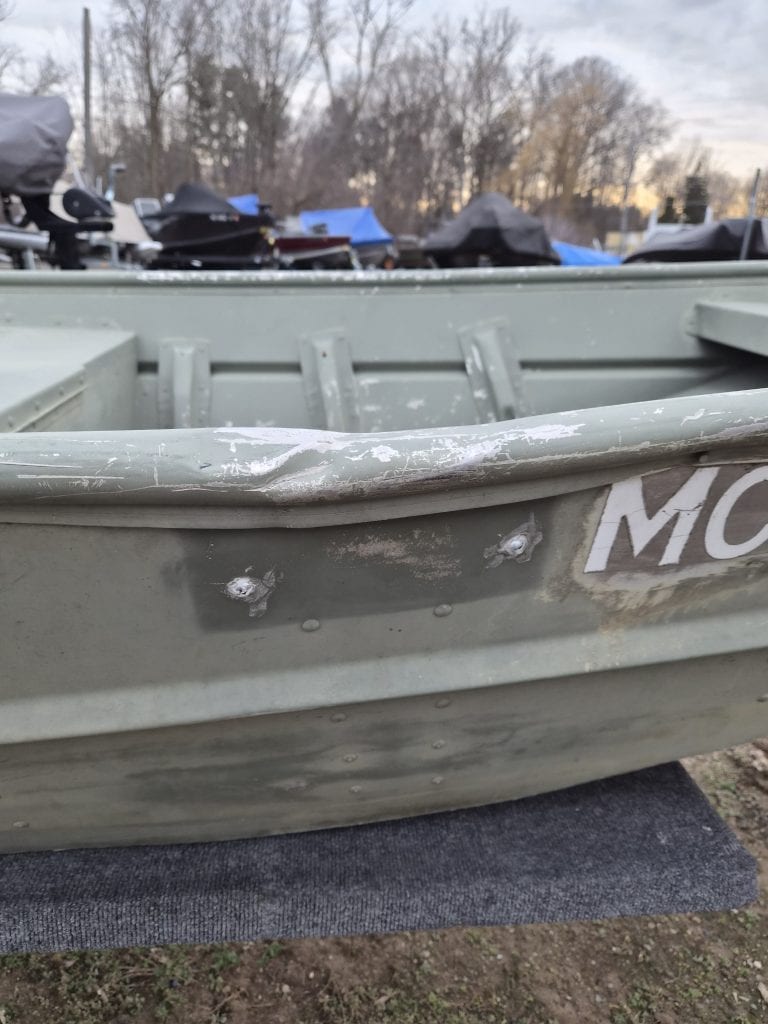 Close-up of a weathered boat hull showing minor damage and repairs, with boat motors and outdoor storage in the background.