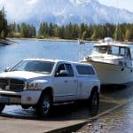 Boat and pickup truck by scenic lake with mountains, trees, and blue sky for outdoor towing and boating.