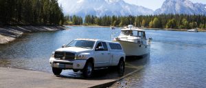 Boat and pickup truck by scenic lake with mountains, trees, and blue sky for outdoor towing and boating.