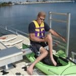 Person entering a bright green kayak at a lakeside docking station with safety gear.