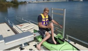 Person entering a bright green kayak at a lakeside docking station with safety gear.