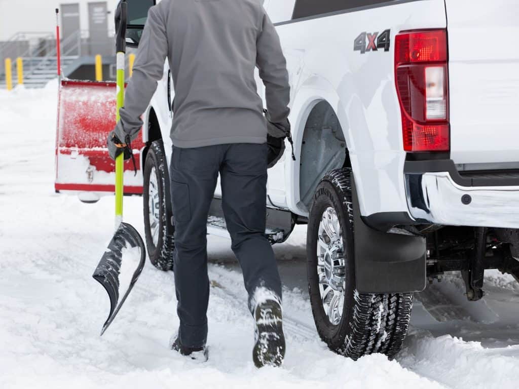 A person clearing snow from a white 4x4 pickup truck with a snow shovel in a snowy parking lot.