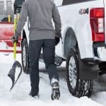 A person clearing snow from a white 4x4 pickup truck with a snow shovel in a snowy parking lot.