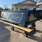 Black truck bed camper shell with windows on a dolly in outdoor lot.