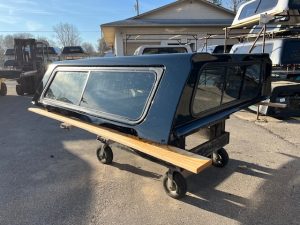 Black truck bed camper shell with windows on a dolly in outdoor lot.