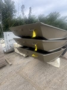 Three brown Jon boats stacked outdoors with trees in background.