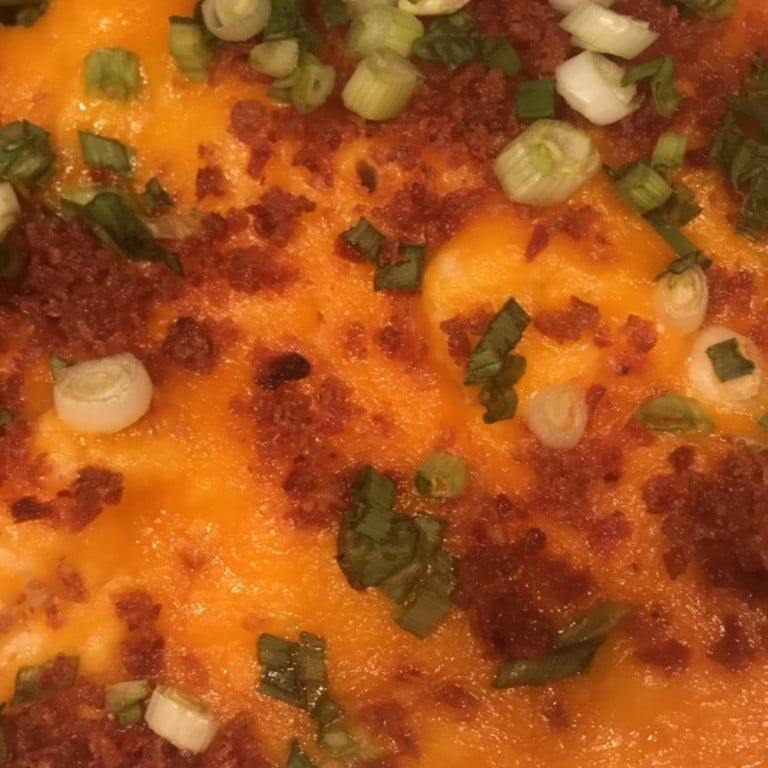 Close-up of loaded mashed potatoes topped with melted cheese, crispy bacon bits, chopped green onions, and seasoning, served as a hearty side dish.