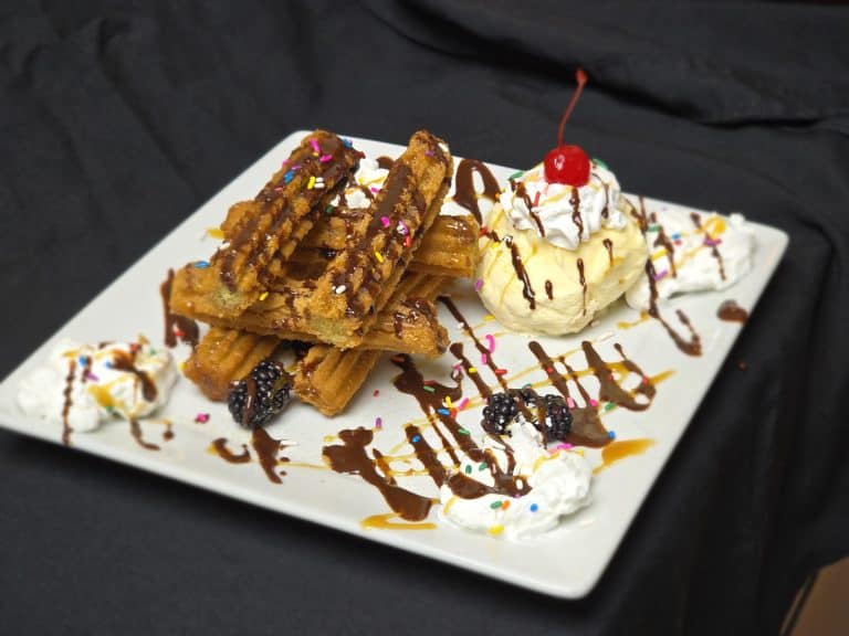 Churros with ice cream, whipped cream, and cherry on a white plate.