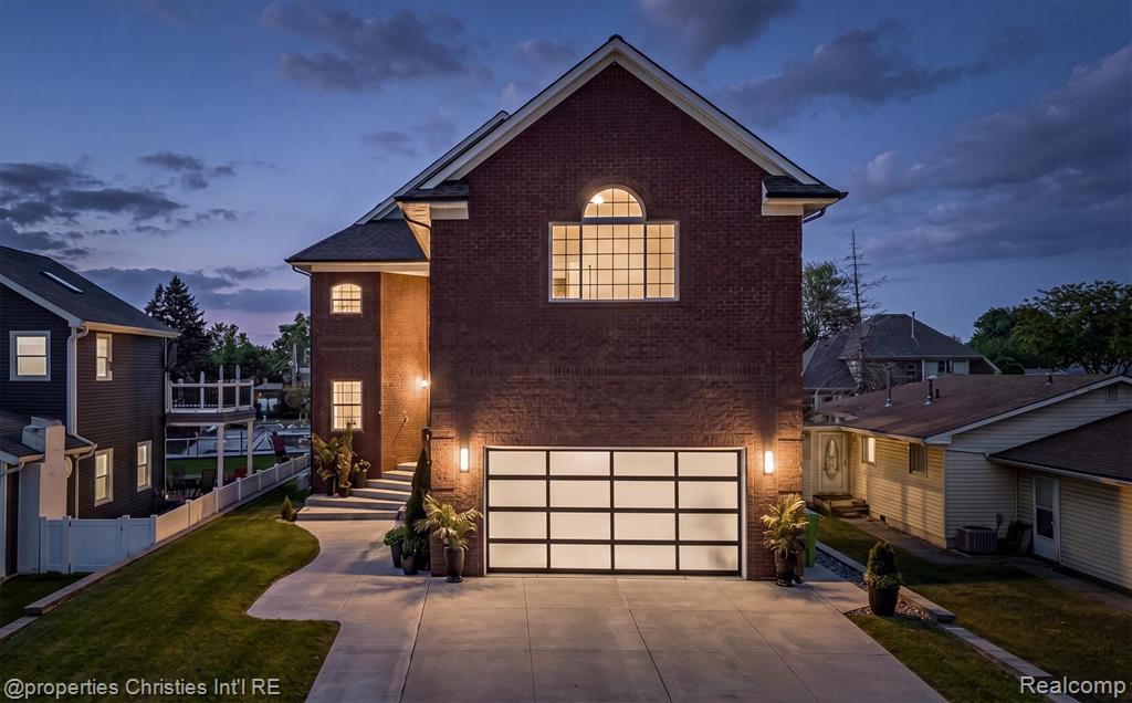 Brick exterior residence featuring an arched window, a modern glass-paneled garage door, concrete driveway, and established landscape plantings