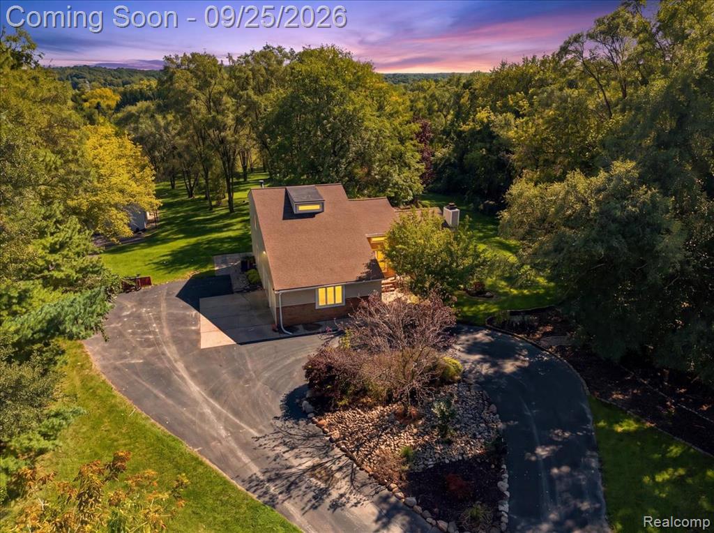 Paved drive, mature trees, landscaped beds, and a brown shingle roof