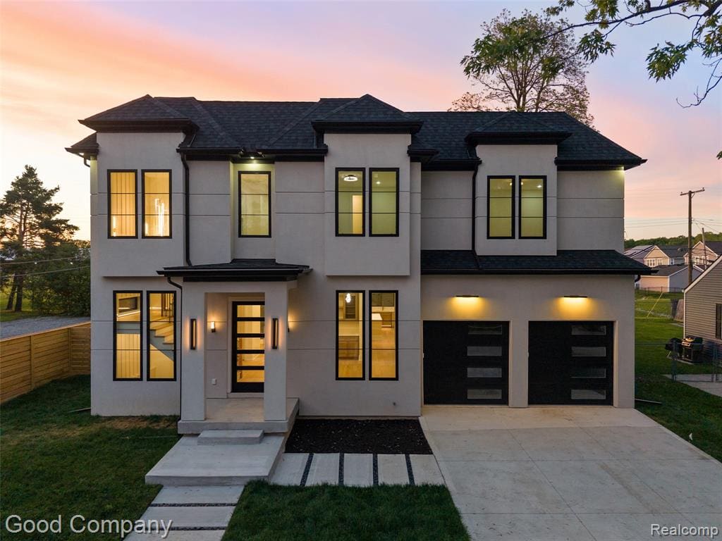 Contemporary two-story residence featuring a light-toned exterior, dark-framed windows, a covered entry, an integrated two-car garage with modern doors, and a concrete driveway