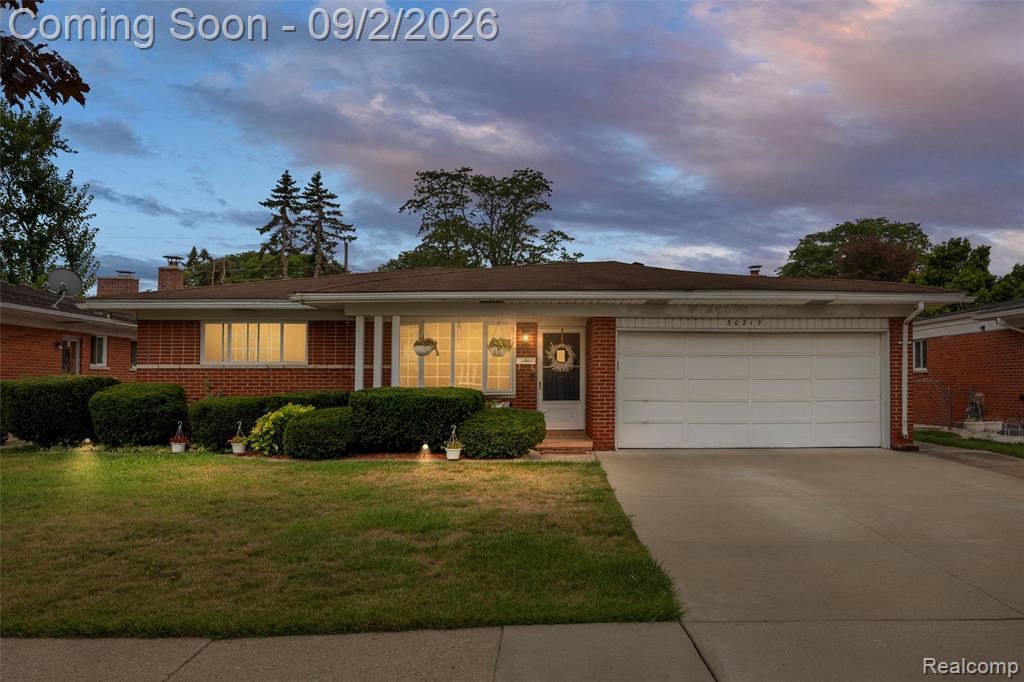 Brick facade ranch home featuring an attached garage, concrete driveway, and mature front landscaping
