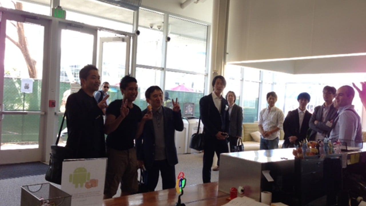 A group of three people posing for a photo inside a building full of windows, making a V sign with their fingers, while others watch from the side
