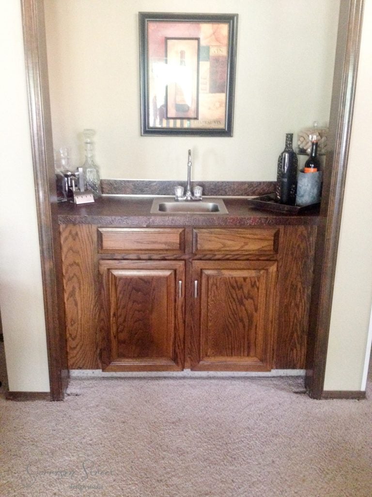 A small bar area with a sink, wooden cabinets, and a countertop. Bottles and glassware are arranged on the counter. A framed abstract art piece hangs above the bar. Carpeted flooring is visible.