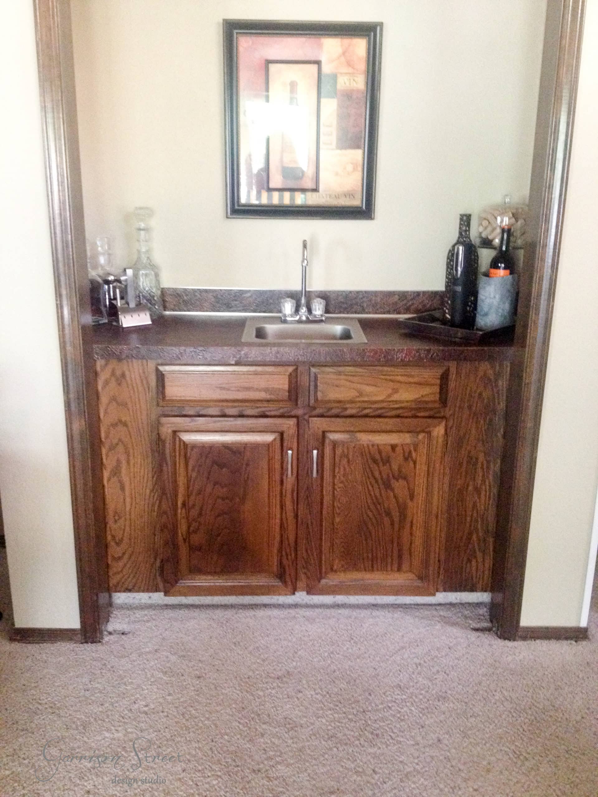 A small bar area with a sink, wooden cabinets, and a countertop. Bottles and glassware are arranged on the counter. A framed abstract art piece hangs above the bar. Carpeted flooring is visible.