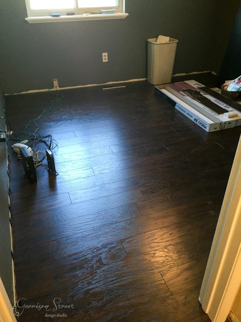 A room with dark wood flooring, gray walls, and a small window features an innovative IKEA desk hack. A white trash bin sits by the wall, surrounded by electrical cords and a modem. Unopened boxes are neatly stacked in the corner, waiting to unveil their secrets.