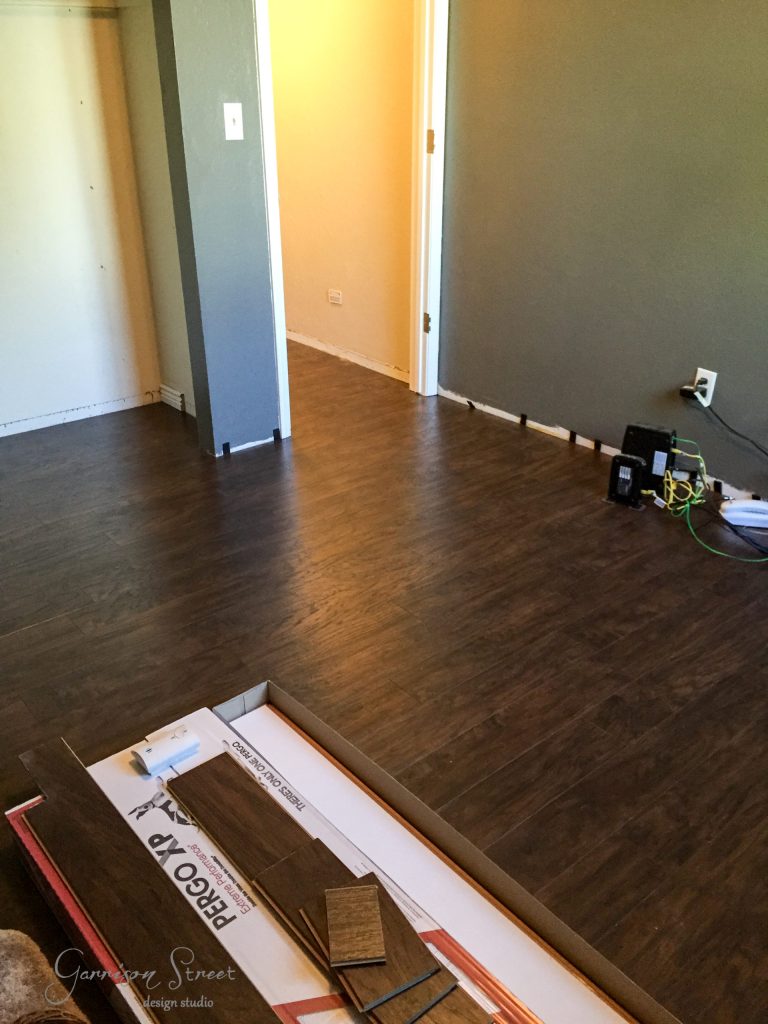 A newly laid dark hardwood floor graces the room, which features light-colored walls and an open closet. Tools and a box with leftover flooring materials hint at recent work, perhaps even an IKEA desk hack in progress. A door leads invitingly to another room.