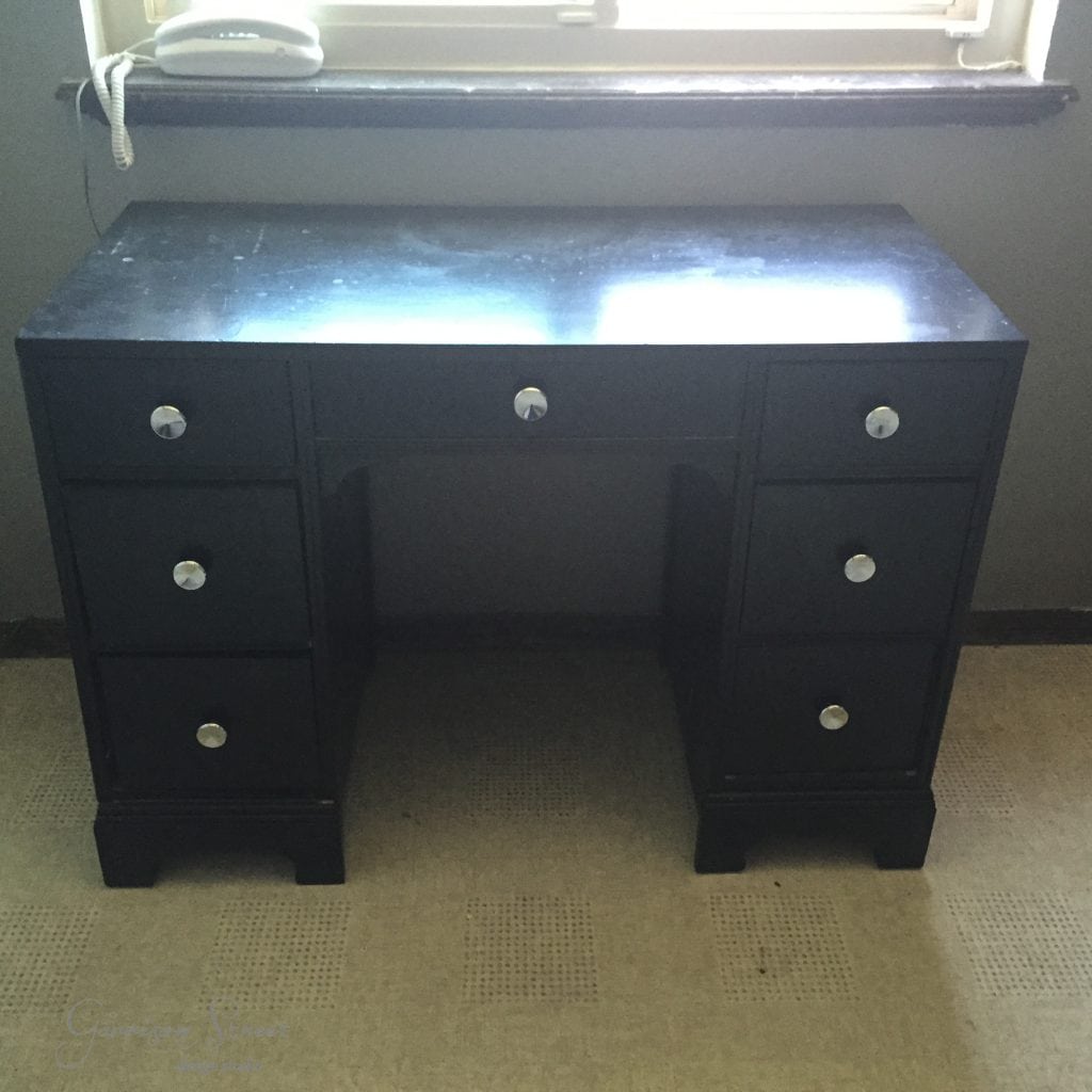 A black wooden desk, reminiscent of an IKEA desk hack, features six drawers with round silver knobs and is placed in front of a window. A white office phone sits to the left, while sunlight partially illuminates its surface.