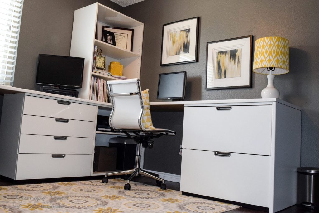 A modern home office features a white desk showcasing an IKEA desk hack, paired with a matching file cabinet and swivel chair. A lamp with a yellow patterned shade sits atop the cabinet, while abstract art adorns the walls. A patterned rug ties it all together over the dark hardwood floor.