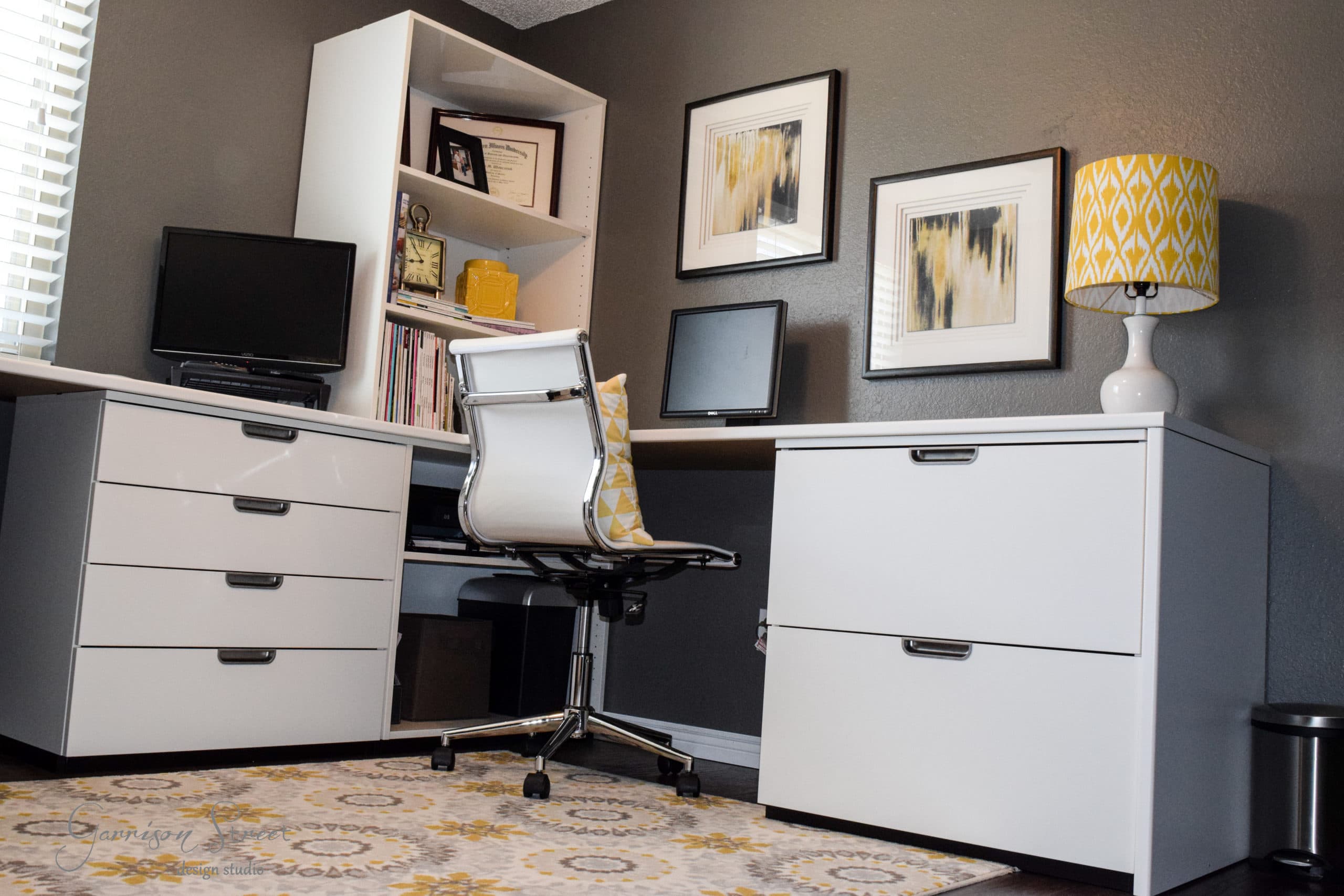 A modern home office features a white desk showcasing an IKEA desk hack, paired with a matching file cabinet and swivel chair. A lamp with a yellow patterned shade sits atop the cabinet, while abstract art adorns the walls. A patterned rug ties it all together over the dark hardwood floor.