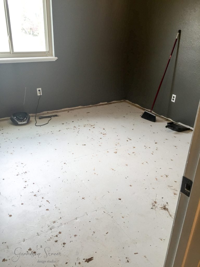 A room with bare, dusty subflooring and gray walls features a creative IKEA desk hack. A broom and dustpan lean against the wall, while a window partially illuminates the space. Nearby, a small device is plugged into the wall near the window, adding a touch of modernity.