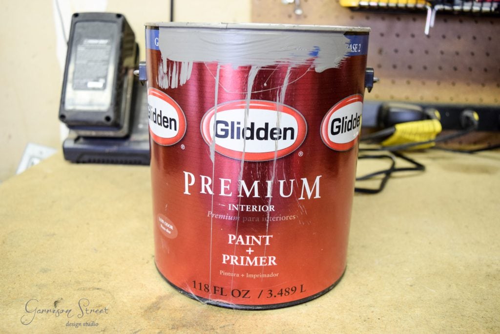 A large red can of Glidden Premium Interior Paint + Primer sits on a workshop table, ready for the next DIY project. Paint drips from the top, hinting at creativity in progress. In the background, an organized pegboard and tools set the scene for an ambitious IKEA desk hack.