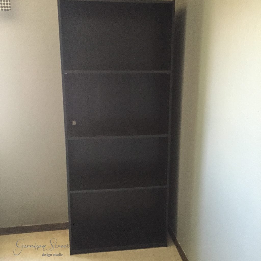 A tall, dark wooden bookcase with four shelves stands proudly in a corner against a light-colored wall, resembling an ingenious IKEA desk hack. The bookcase is empty, resting on a floor of light-colored wood.