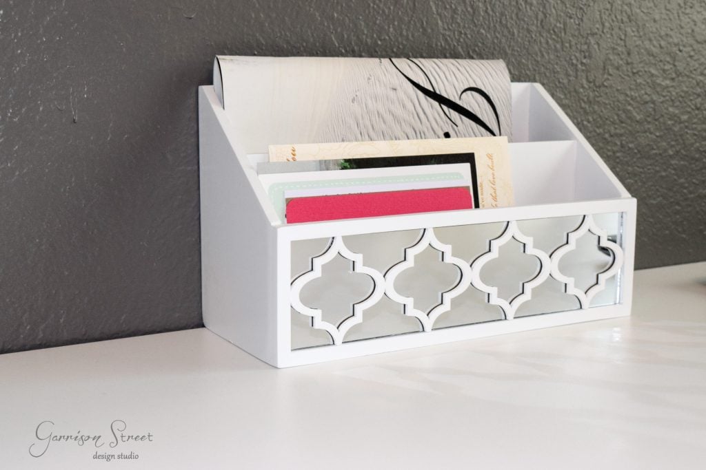 A white decorative letter holder, part of an IKEA desk hack, sits elegantly on a white surface, containing assorted letters and envelopes. The holder boasts a geometric cutout design on the front and is set against a gray wall.