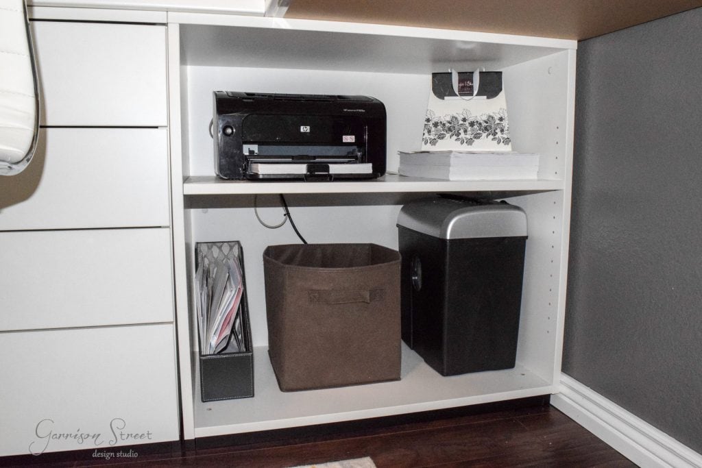 A white shelving unit, reminiscent of an IKEA desk hack, features a printer and a stack of paper on the top shelf. The bottom shelf accommodates a brown fabric bin, a paper shredder, and vertical file organizers with documents. This tidy ensemble complements the home office's hardwood floor.