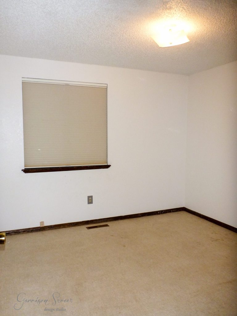 An empty room with beige walls and carpet features a single closed window with a shade. The ceiling boasts a bright light fixture, and visible baseboard trim lines the walls—perfect for an IKEA desk hack to transform this blank slate into an inspiring workspace.