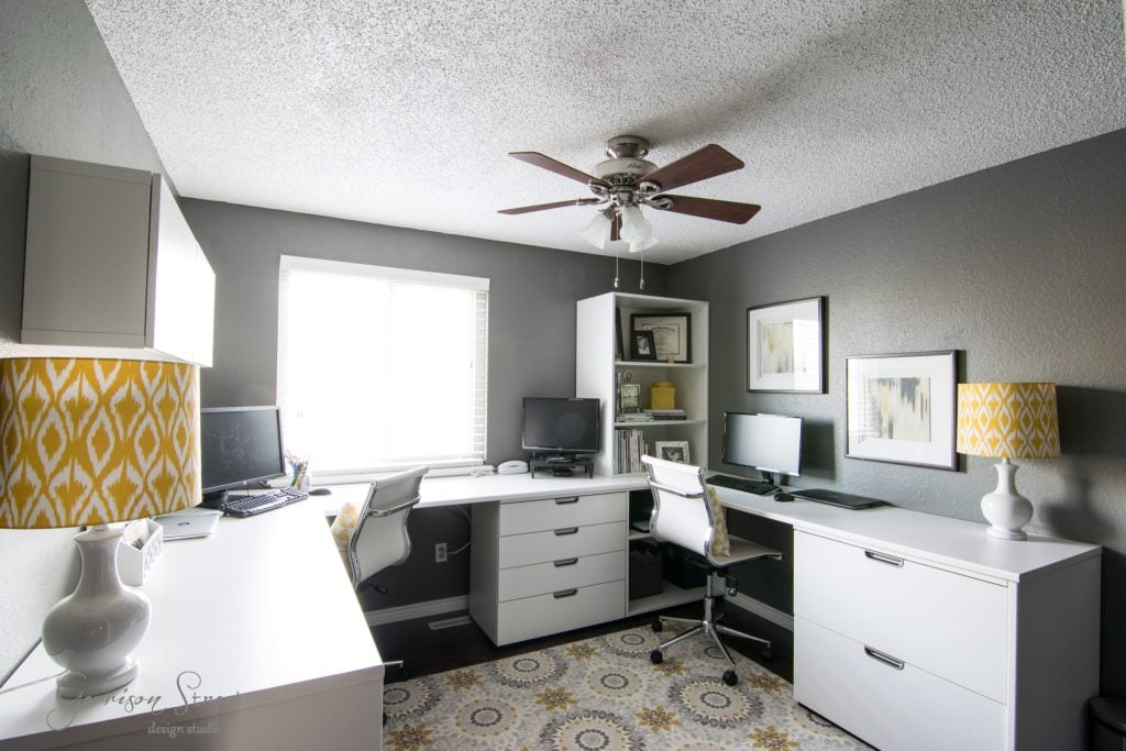A modern home office with an IKEA desk hack showcases L-shaped white desks, two computer monitors, a white office chair, and yellow patterned lamps. Gray walls feature framed art, and a window with blinds allows natural light. A ceiling fan is above, and a patterned rug adorns the floor.