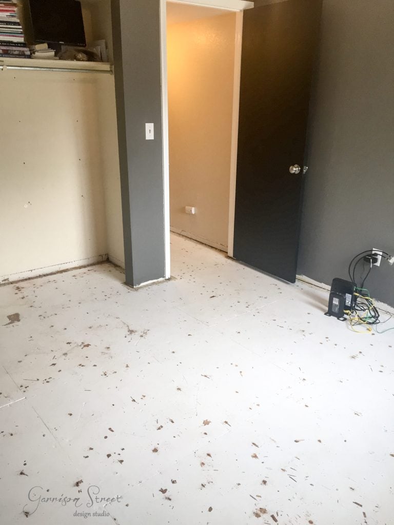 A partially renovated room with a bare, speckled floor and walls painted gray and beige. An open doorway contrasts a closed black door. Cables and electronic devices clutter the corner, while an IKEA desk hack adds functional style to the evolving space.