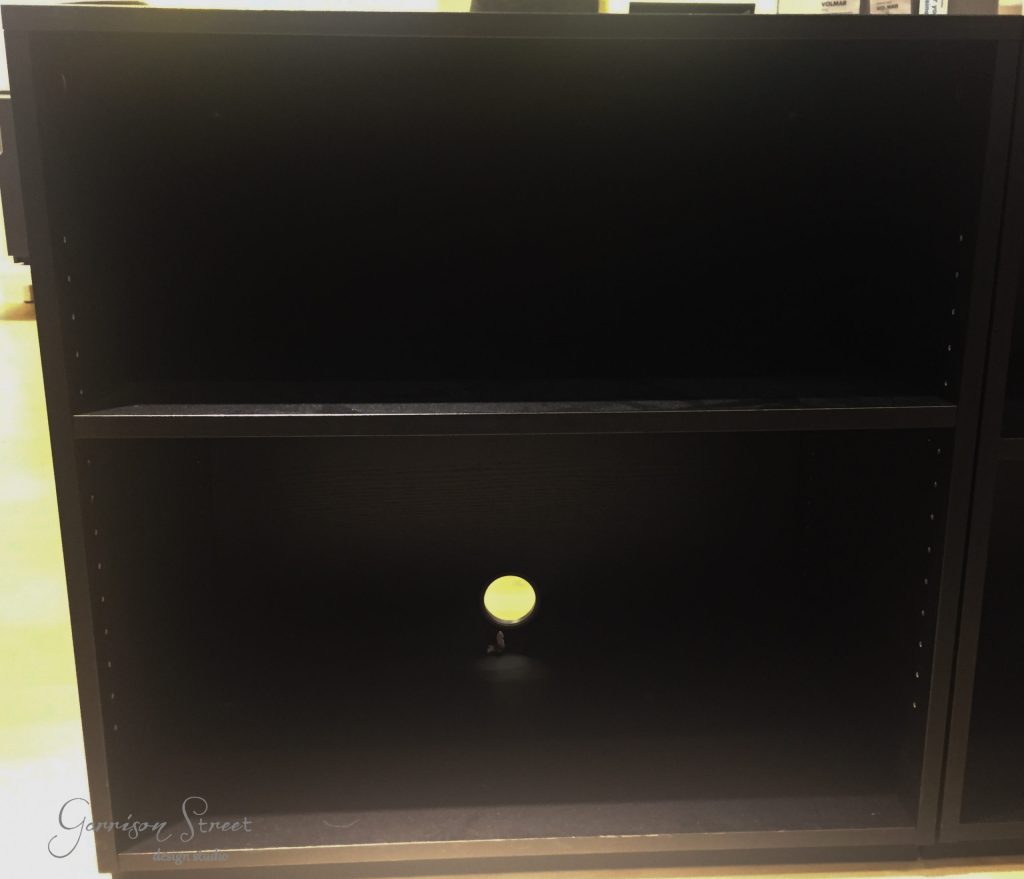 A black empty shelving unit, perfect for an IKEA Desk Hack, features two horizontal compartments and a circular hole on the lower shelf for seamless cable management. Positioned indoors on a light-colored floor, it seamlessly blends functionality with minimalist design.