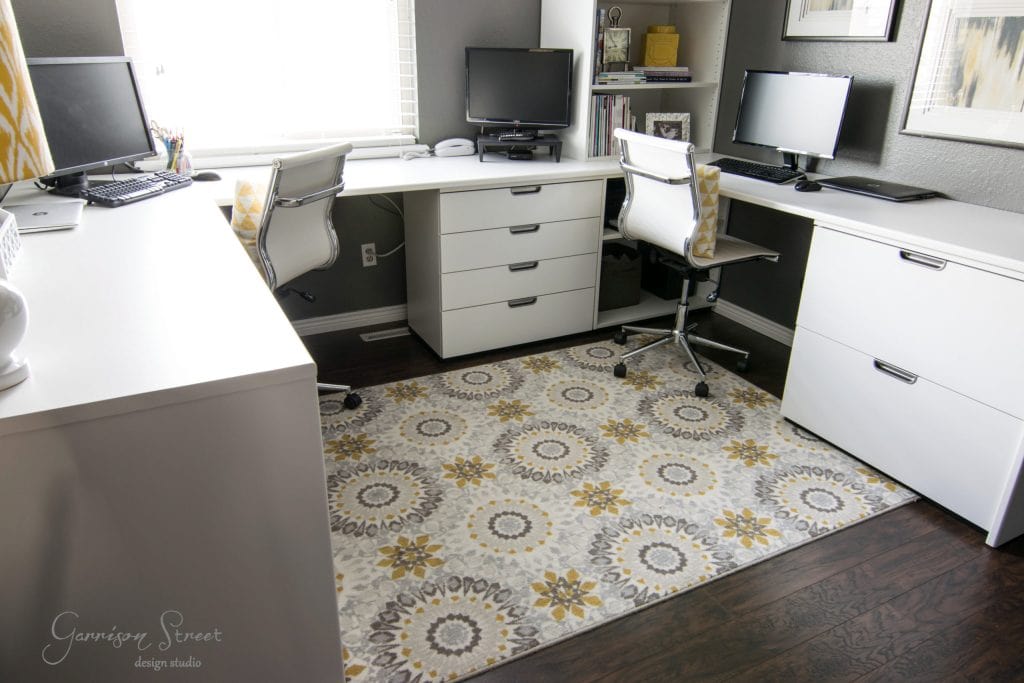 A modern home office features an innovative IKEA desk hack, with two desks boasting sleek monitors and chic white chairs. Light gray walls, a large window, and a patterned rug with yellow and gray geometric designs on dark wooden floors complete the look.