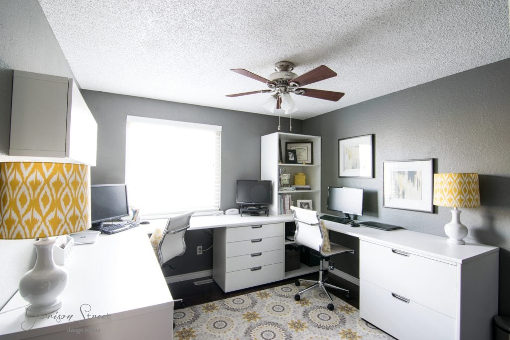 A modern home office with white furniture and gray walls features a U-shaped IKEA desk hack arrangement, dual monitors, white chairs, and decorative lamps with yellow-patterned shades. A ceiling fan and window add light and airiness to the space.