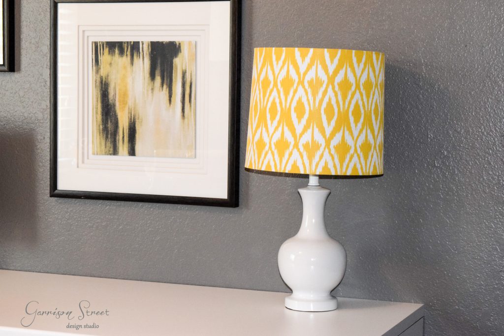 A white lamp with a yellow patterned shade sits on an elegantly hacked IKEA desk against a gray wall. Above the desk, a framed abstract painting with black and yellow brushstrokes adds a creative touch to the setup.