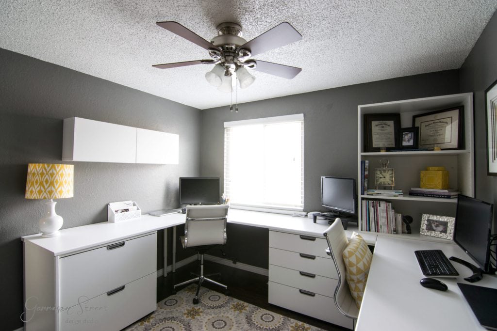 A modern home office features a stylish IKEA desk hack with a white L-shaped design, two chairs, a computer, and a laptop. Shelving units display books and decor. A ceiling fan is perfectly centered above, while a large window floods the space with natural light against gray walls and a patterned rug.