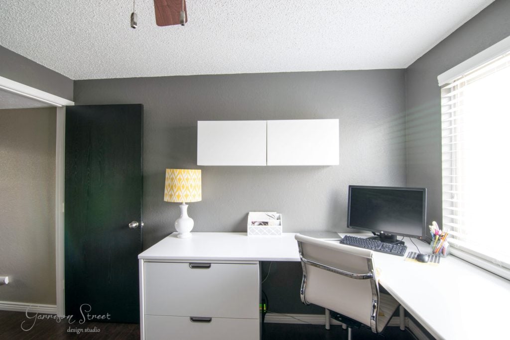 A modern home office features an IKEA desk hack with a white desk, computer, and chair. A yellow and white lamp rests atop a filing cabinet, while wall-mounted cabinets offer storage. The gray walls bask in sunlight streaming through a large window.