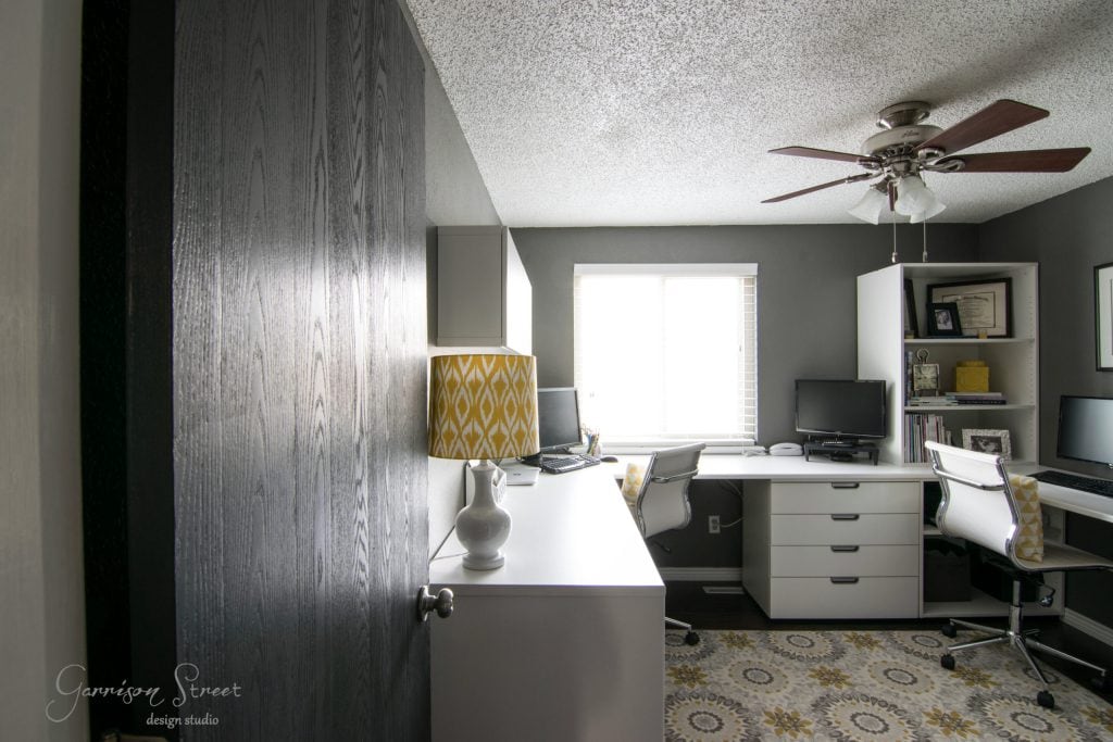 A modern home office features a dark wooden sliding door and boasts an IKEA desk hack with white desks perfectly arranged under a bright window. A ceiling fan with lights hangs overhead, while a patterned rug adds charm to the floor.