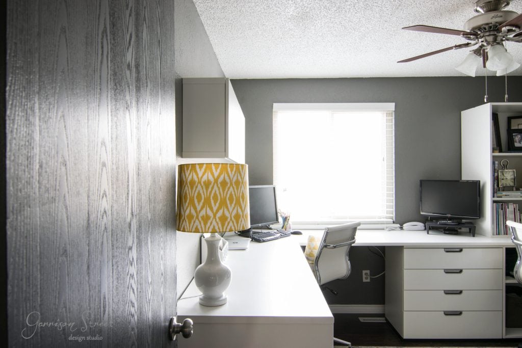 A modern home office with gray walls showcases an IKEA desk hack setup. A yellow-patterned lamp graces the left side, accompanied by two swivel chairs and a desktop computer. Storage cabinets provide organization, while a ceiling fan spins above. Natural light streams through the window, completing the space. .