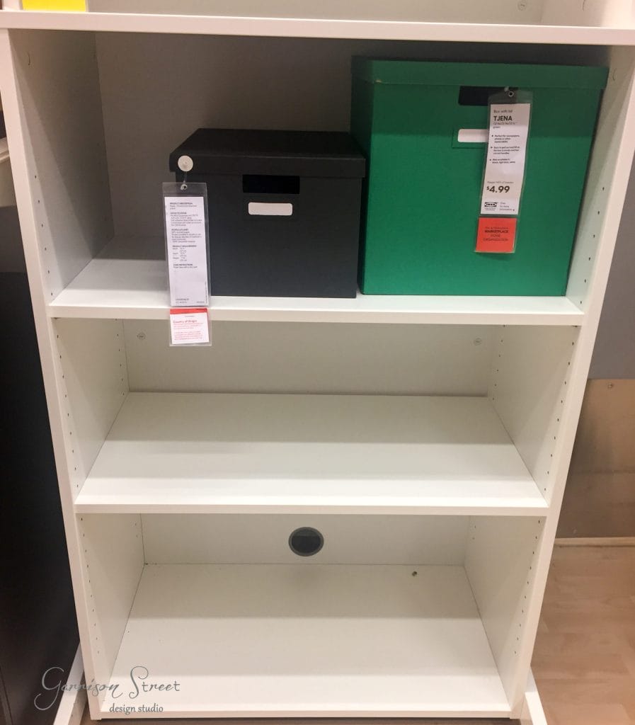A white, three-shelf storage unit, reminiscent of an IKEA desk hack, showcases a black box with a label on the top shelf and a green box with a label on the middle shelf. The bottom shelf remains empty. The open-backed design features a circular cutout for seamless cord management.