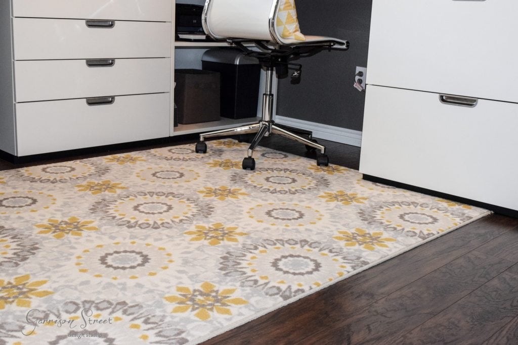 A white and gray office chair on wheels sits on a patterned rug with floral motifs and geometric designs in yellow, gray, and white. The chair is positioned between white drawers on a wooden floor, perfectly complementing an IKEA desk hack that adds both style and function to the workspace.