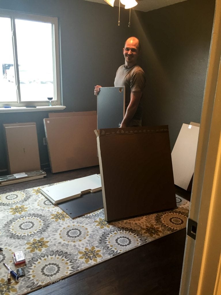 A person is in a room with several pieces of wood and packing materials on the floor, likely assembling an IKEA desk hack. The room has dark walls, a window, and a patterned rug.