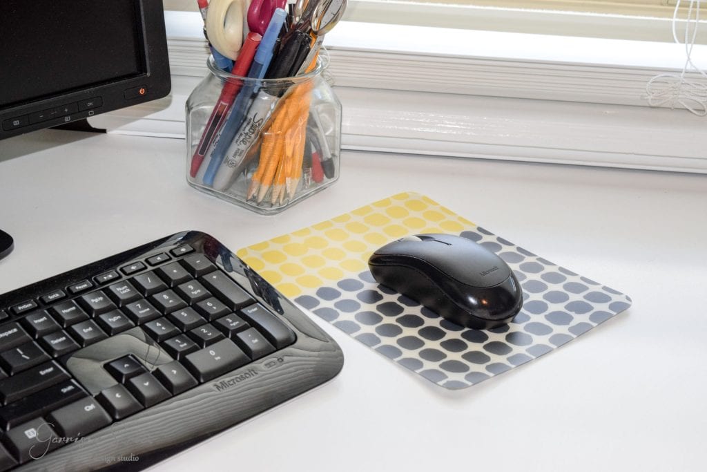 A computer keyboard and mouse rest on a white IKEA desk. The mouse is on a pad with a yellow and gray polka dot pattern. Nearby, a glass jar holds assorted pens and colored pencils, showcasing an ingenious IKEA desk hack. A monitor is partially visible in the top left corner.