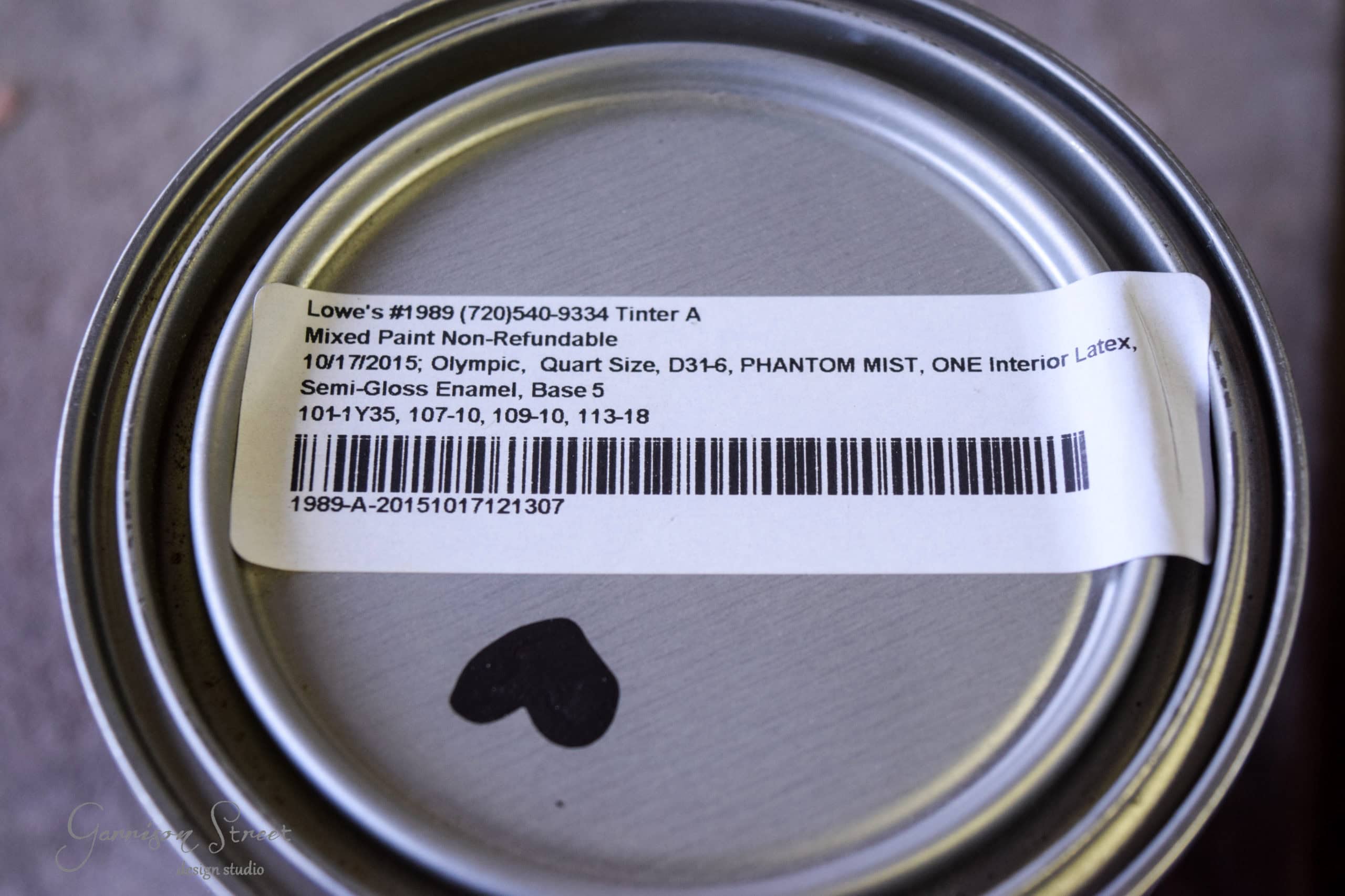 A close-up of a paint can lid with a label from Lowe's. The label details a mixed paint, non-refundable, with specific tint information: Olympic, Quart Size, Phantom Mist, Semi-Gloss Enamel, Base 5. It also includes a barcode and numerical codes.