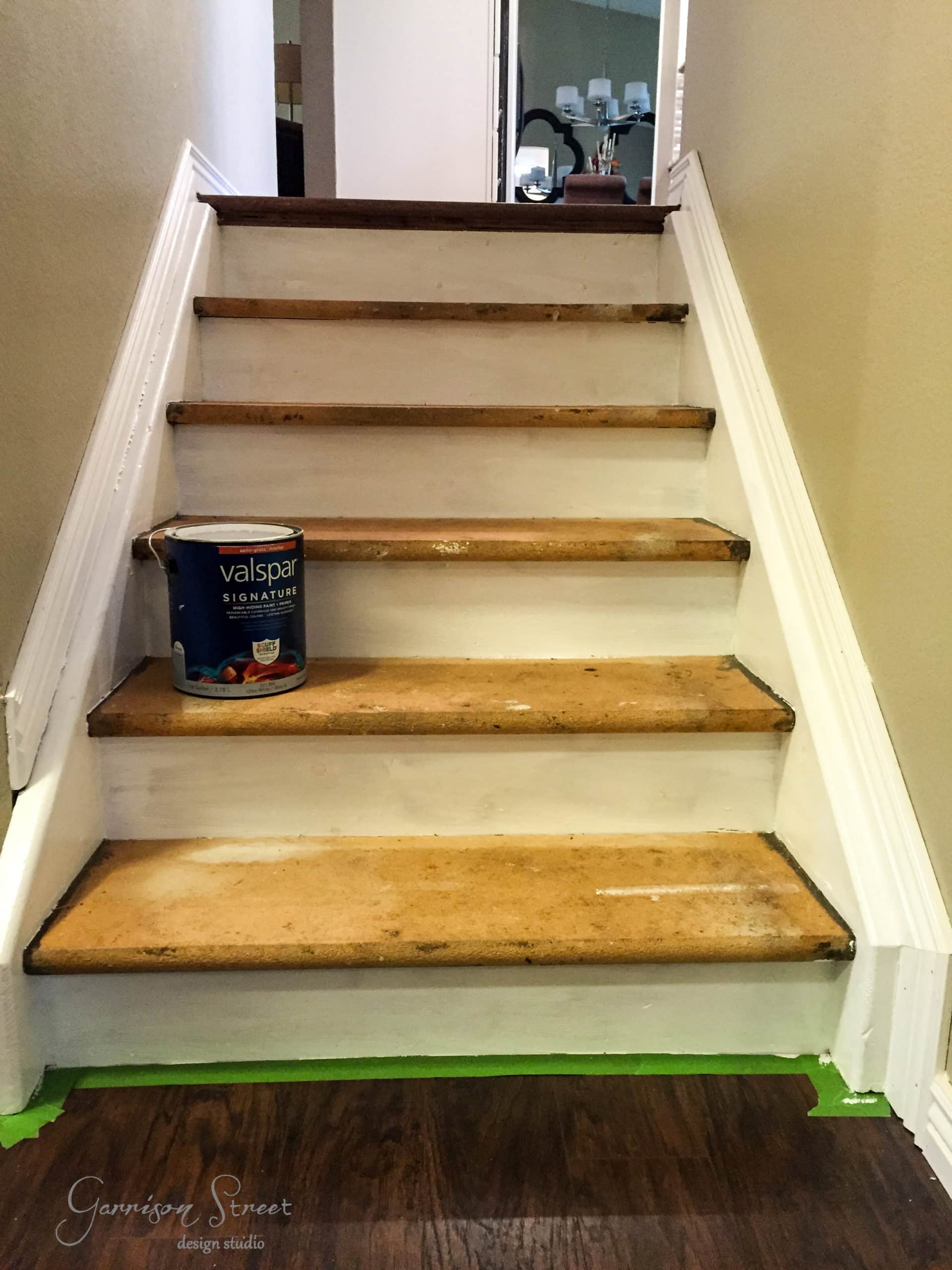 Staircase renovation in progress with wooden steps partly sanded. A can of Valspar paint sits on one step, while protective tape lines the floor at the base.