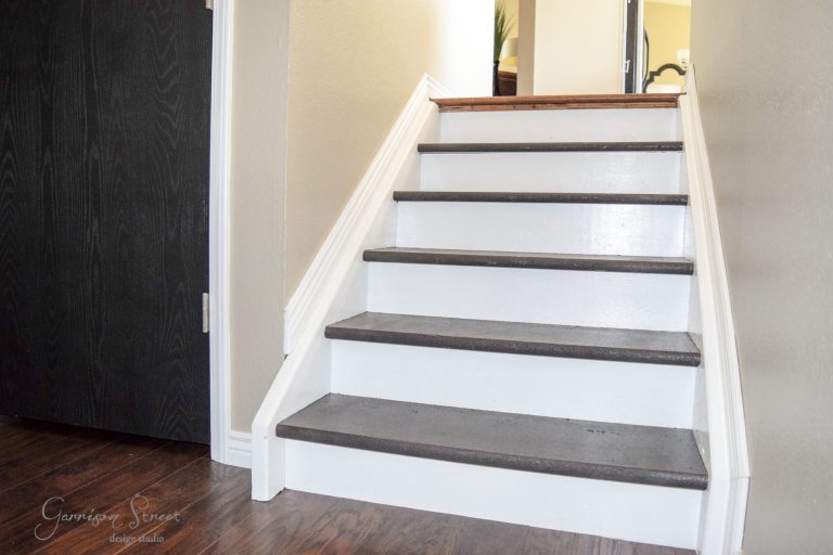 A small staircase with dark wood treads and white risers leading up to a hallway. The stairs are bordered by a beige wall and dark door on the left. The bottom of the stairs is adjacent to a dark wooden floor.