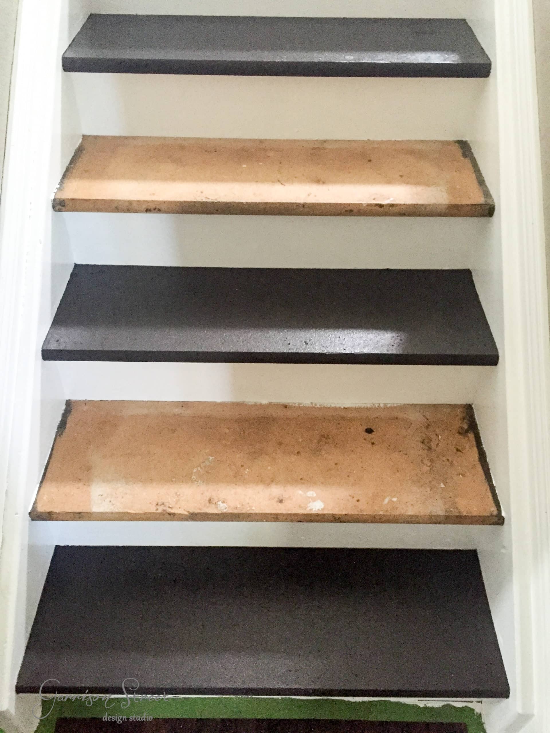 A staircase with alternating wood and dark painted steps. The wood steps appear worn, while the dark steps are smooth and freshly painted. The surrounding walls are white, creating a contrast with the staircase.