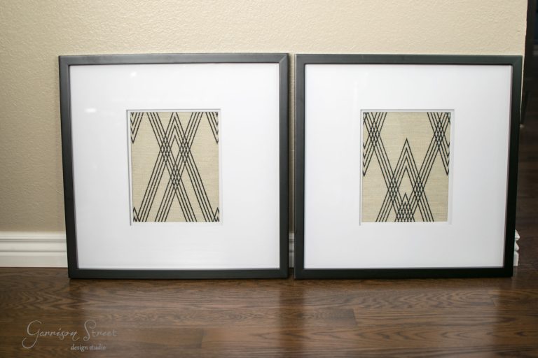 Two framed artworks rest on the wooden floor, each showcasing a symmetrical geometric design with intersecting black lines on a beige background. The frames are black with a white mat, lending the pieces a DIY charm reminiscent of intricate fabric art.