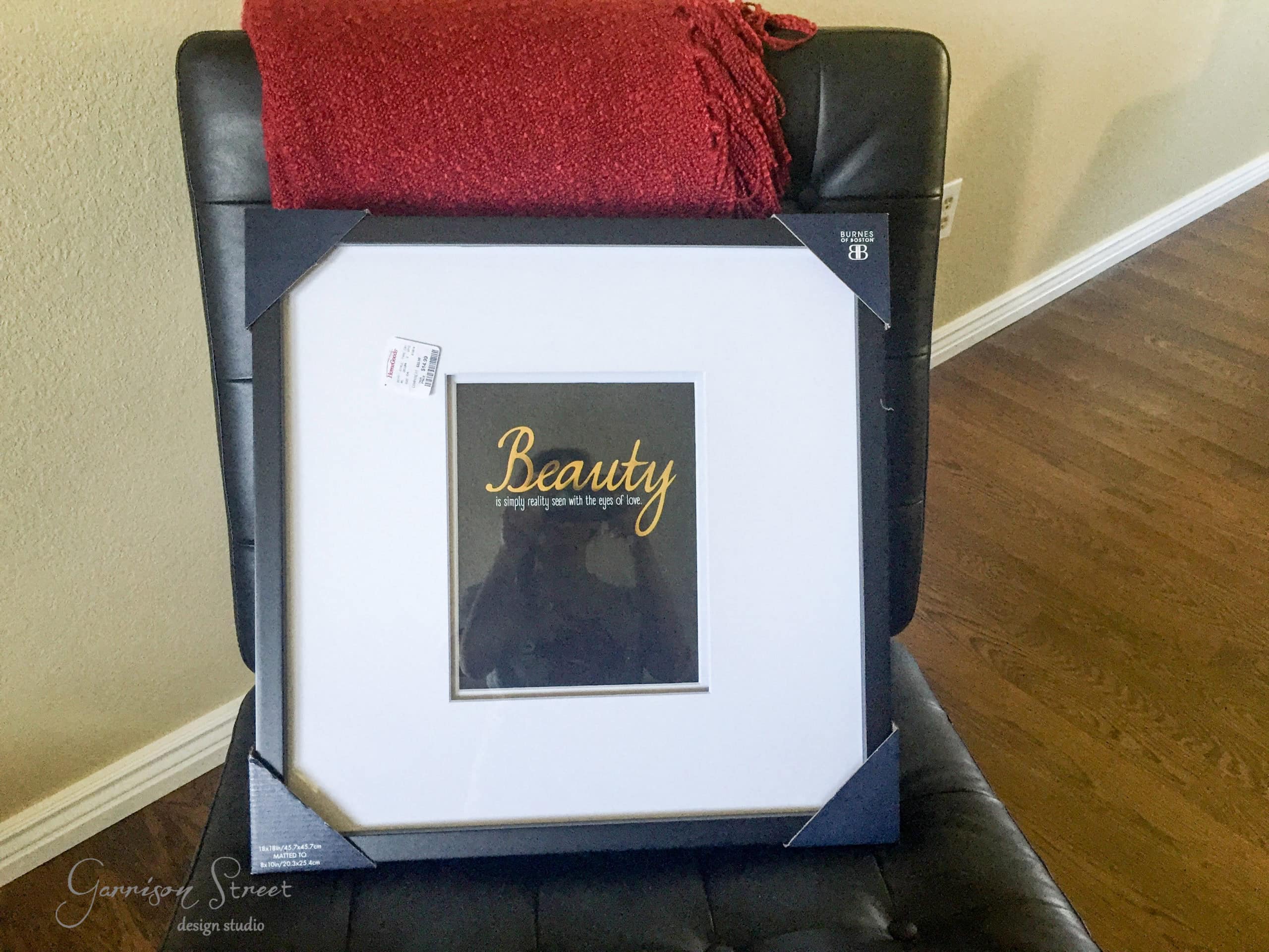 A framed picture with "Beauty" in gold on a black background sits on a black chair, adorned with a red knitted blanket. This piece of DIY fabric art rests elegantly on the wooden floor against a plain wall, creating a striking contrast.